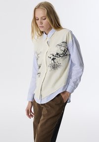 Camicia a bottoni con un motivo a righe blu e bianche indossata sotto un gilet color crema con un'illustrazione nera di elementi naturali. Tessuto testurizzato. Pantaloni marroni.