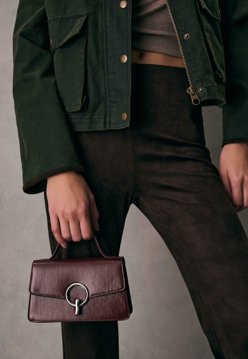 Personne portant un pantalon foncé et une veste verte, tenant à côté d'elle un petit sac à main en cuir marron avec un fermoir circulaire argenté.