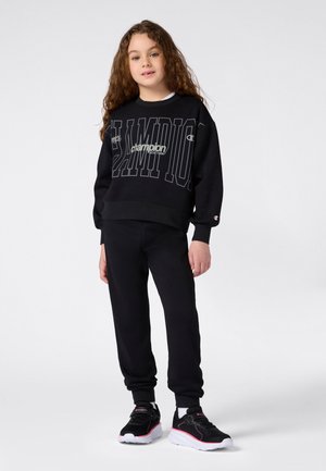 Joven con cabello largo y rizado viste un sudadera negra de Champion, pantalones de chándal a juego y zapatillas negras con suelas blancas y acentos rosas.