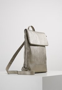 Hopeapintainen nahkareppu, jossa on läppäkiinnitys, säädettävä olkahihna ja ylhäällä oleva kantokahva, levätessään valkoisella pinnalla yksinkertaista taustaa vasten.