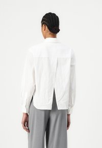 Camicia bianca a maniche lunghe con uno spacco sul retro, realizzata in un tessuto testurizzato. Presenta polsini con bottoni e un colletto a punta. Indossata con pantaloni grigi.