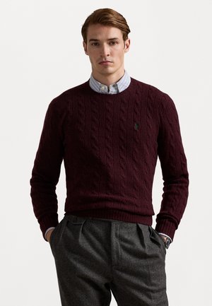 Burgunderfarbene Kabelstrickjacke mit Rundhalsausschnitt und gerippten Bündchen, kombiniert mit grauen hochgeschnittenen Hosen. Dezentes Logo auf der Brust.