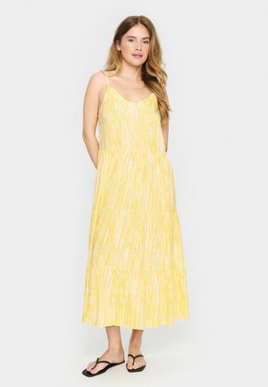 Gelbes Maxikleid mit dünnen Trägern. Verfügt über ein gestuftes Design und ein lineares weißes Muster. Getragen mit schwarzen Flip-Flops.
