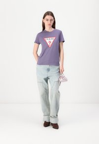 Lilla bomulls t-skjorte med en rød og hvit trekantet logo, sammen med lyseblå baggy jeans og brune sko. Holder en liten lavendel veske.