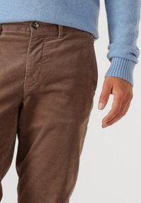 Man wearing brown corduroy pants and a light blue sweater with ribbed cuffs, hand relaxed at side against a neutral background.