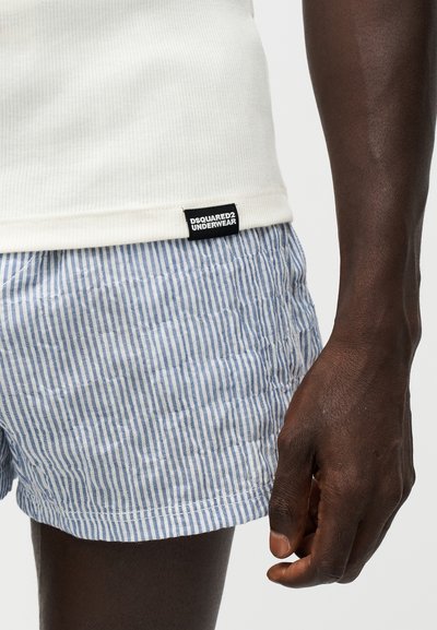 Gros plan d'une personne portant un short rayé bleu et blanc et une chemise blanche avec une étiquette visible "Dsquared2 Underwear".