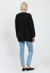 Schwarzer Strickpullover mit lockerer Passform und langen Ärmeln, der kleine strukturierte Details aufweist; getragen mit hellblauen Jeans und schwarzen Schuhen.