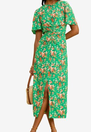 Vrouw draagt een groene bloemen midi-jurk met wapperende mouwen en een split aan de voorkant, en houdt een gevlochten strohandtas met leren bandjes vast.