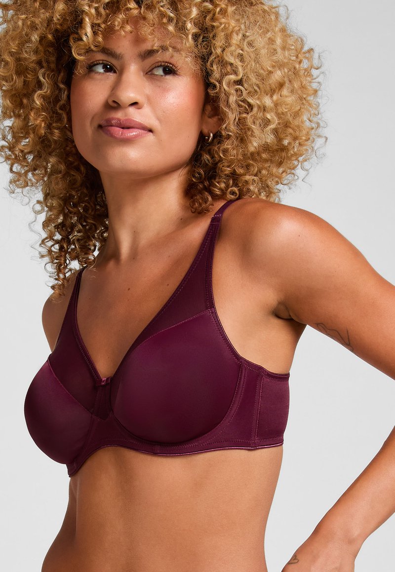 Woman with curly hair wearing a dark purple supportive bra, looking to the side with a neutral expression against a plain gray background.