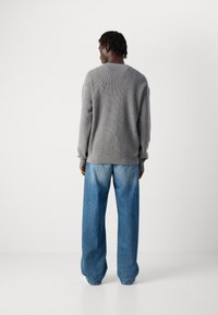 Mann trägt einen grauen Strickpullover und lockere, hellblaue Jeans, steht mit dem Rücken zu einer weißen Wand.