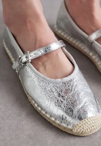 Silver metallic espadrille flats with a strap, featuring a textured upper, rounded toe, and a natural jute sole.
