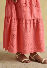 Rochie maxi din bumbac corai, cu broderii intricate și modele decupate, având un design în straturi și un accent la tiv. Purta cu sandale bej.