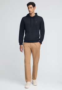 Marineblå hoodie med framficka och dragsko, tillsammans med beiga chinos och vita sneakers. Enkel, avslappnad design, bomullsmaterial.
