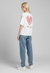 Balta per didelė t-shirt su dideliu raudonu širdies grafiku ant nugaros ir tekstu "SWEET heart", priekyje papuošta spalvingais gėlėmis.