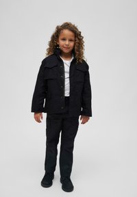 Veste en coton noir avec deux poches frontales, associée à un pantalon noir assorti et des chaussures noires, offrant une coupe décontractée et une fermeture à boutons.