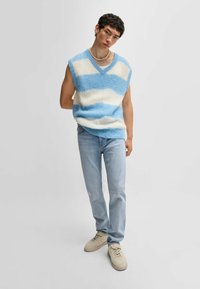 Gilet a righe azzurro e bianco, senza maniche, in maglia con scollatura a V. Indossato su jeans di denim azzurro chiaro e sneakers beige. Design semplice e texturizzato.