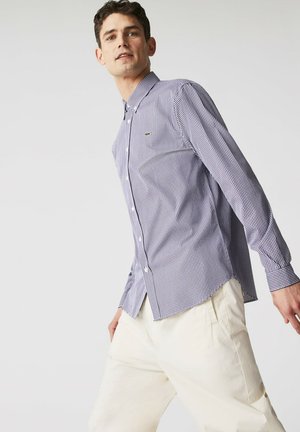 Camisa de botones hecha de tejido tejido, con un patrón de cuadros en púrpura y blanco, de manga larga, y un pequeño logo bordado en el pecho.