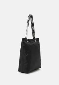 Sac fourre-tout noir avec une texture douce, de forme rectangulaire, et une large sangle portant la marque répétée "PUMA" en blanc.