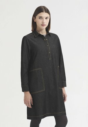 Jeune femme aux cheveux bruns et raides porte une robe en denim foncé longueur genou avec poches avant et col boutonné, debout devant un fond uni.