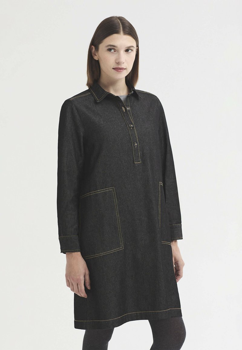 Jeune femme aux cheveux bruns et raides porte une robe en denim foncé longueur genou avec poches avant et col boutonné, debout devant un fond uni.