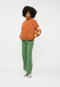 Hnedý fleecový pulóver s polovičným zipsom a bielou kvetinovou výšivkou na rukáve. Skombinovaný so zelenými kordovými nohavicami. Bielé topánky.