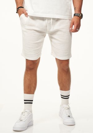 Weiße gewobene Shorts mit elastischem Bund und Kordelzug. Kombiniert mit weißen Sneakers und gestreiften Knöchelsocken. Die Textur wirkt leicht.