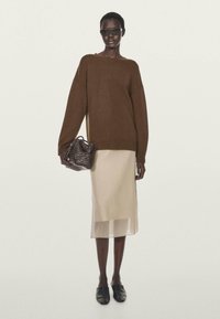 Donna che indossa un maglione marrone oversize, una gonna midi beige, scarpe nere slip-on, occhiali da sole e porta una borsa intrecciata marrone.