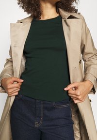 Beige trenchcoat över en mörkgrön topp med slät yta och rund halsringning. Mörkblå jeans med gula sömmar är synliga.