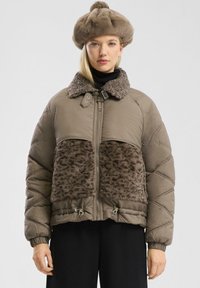 Młoda kobieta w brązowej kurtce puchowej z panelami i kołnierzem z sztucznego futra, pasującym futrzanym kapeluszu i czarnych spodniach na jednolitym tle.