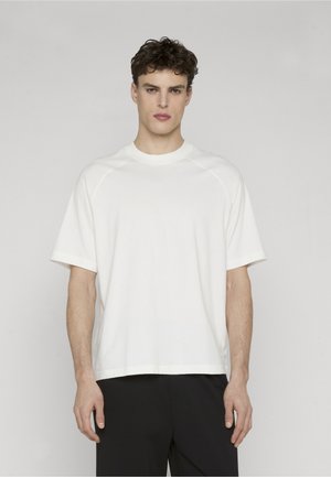 Jonge man met krullend haar, gekleed in een effen wit T-shirt met korte mouwen en zwarte broek, staand met een neutrale uitdrukking tegen een grijze achtergrond.