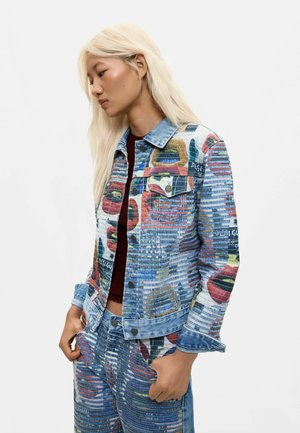 Junge Frau mit langen blonden Haaren, die eine farbenfrohe gemusterte Jeansjacke und passende Jeans trägt, steht mit den Händen in den Taschen.