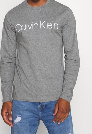 Mężczyzna w szarej koszuli z długim rękawem z białym napisem „Calvin Klein” oraz w niebieskich, przecieranych dżinsach.