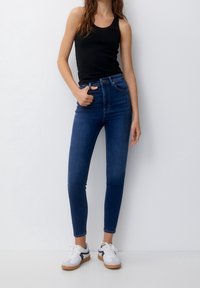 Un haut sans manches noir associé à un jeans skinny bleu en denim taille haute. Le mannequin porte des baskets blanches avec des accents noirs. Design simple, texture lisse.