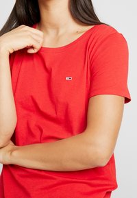 Röd bomull t-shirt med rund halsringning. Kort ärm och en liten logotyp med röda, vita och blå accenter nära bröstet. Slät textur.