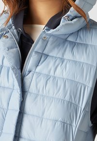 Ljusblå pufferväst, isolerad, med horisontellt quiltat mönster, dragkedjestängning och huva, tillverkad av smidigt, vattenavvisande material.