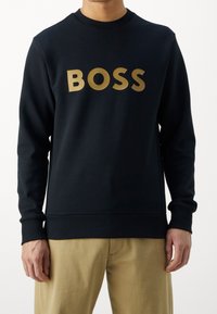 Persona vestida con sudadera negra con el texto dorado "BOSS" y pantalones beige, mostrada desde el cuello hasta la mitad del muslo sobre un fondo blanco.