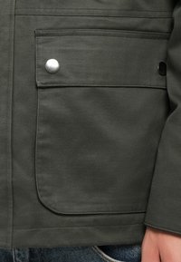Donkergroene jackedetail met een grote zak en een zilveren drukknoop. Het materiaal lijkt glad met een matte afwerking.
