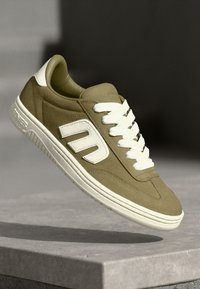 Olivově zelené tenisky s bílými akcenty, s texturovaným semišovým svrškem, bílými tkaničkami a silnou gumovou podrážkou. Viditelné logo na boku.