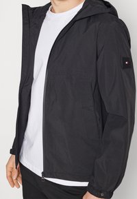 Mand iført svart hættetrøje med lynlås over hvid t-shirt, med lynlåslommer og knappede manchetter, vist fra midt på brystet til hofterne.