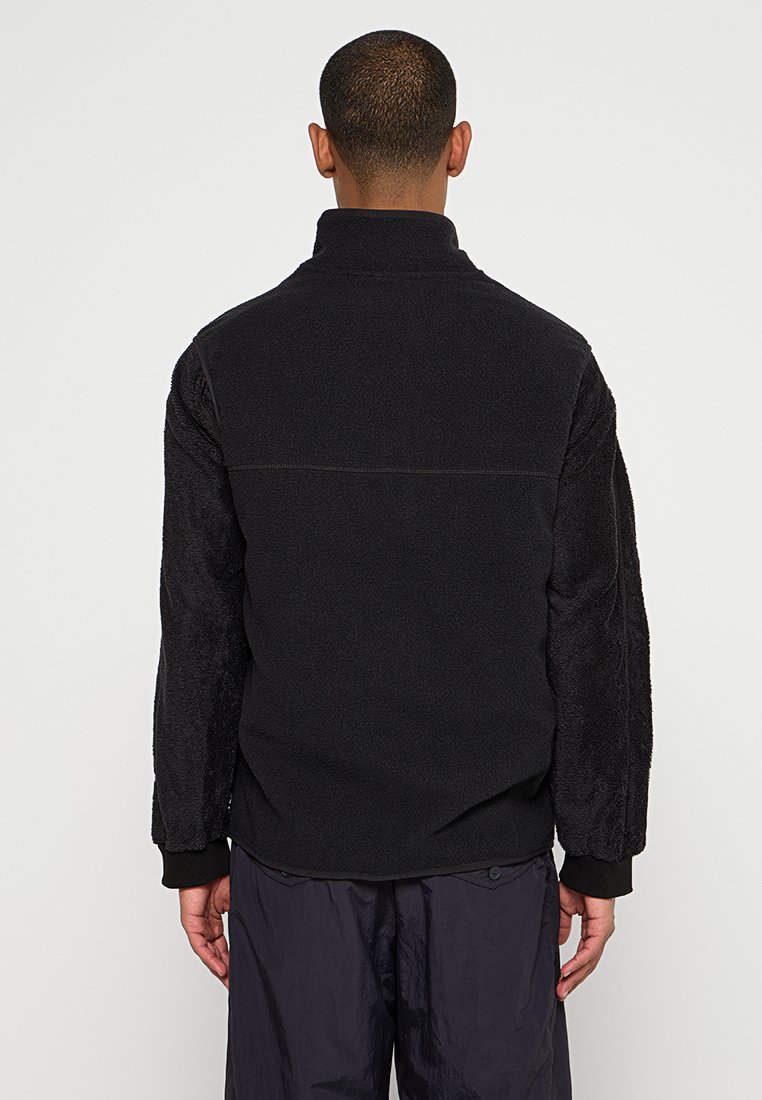 Veste en polaire noire avec col zippé, présentant des manches texturées et un corps lisse et élégant. Poignets élastiques et coupe décontractée.