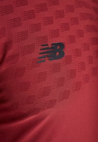 Röd sporttröja med ett texturerat mönster och en svart New Balance-logotyp. Tyget verkar vara lätt och andningsbart.
