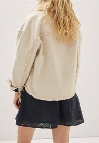 Beige oversized jakke med frynser, båret over en sort broderet kjole med blomsterudskæringer og en let, flydende tekstur.