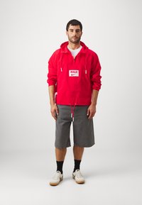 Röd lättviktsjacka med dragkedja, framfickor, dragsko i nederkant och "BULLS"-logotyp. Kombinerad med grå shorts och vita sneakers.