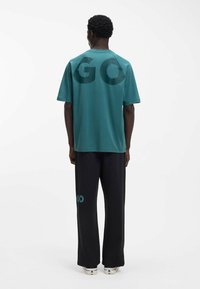 Krótka koszulka z krótkim rękawem w kolorze zielonym morskim, z dużym ciemnym napisem „GO” na plecach, zestawiona z czarnymi spodniami z akcentem „GO” w kolorze zielonym morskim. Gładka tkanina o luźnym kroju.