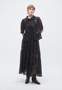 Μαύρο σύνολο δύο τεμαχίων από δαντέλα που περιλαμβάνει μπλούζα με κοντά μανίκια και κουμπιά, καθώς και μακριά φούστα με στρωματωτές βολάν, συνδυασμένο με μαύρα μπότες με τακούνι.