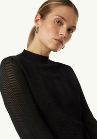Schwarzer Strickpullover mit hohem Kragen, der ein strukturiertes Zickzackmuster und lange Ärmel aufweist. Glatte Oberfläche mit gerippten Akzenten.