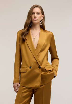Donna con capelli lunghi castani che indossa un completo doppiopetto in satin color senape, orecchini dorati e una collana con pendente, in piedi davanti a uno sfondo uniforme.