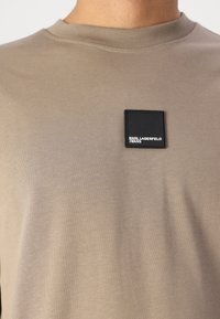 Šviesiai rudas trumpomis rankovėmis megztinis pagamintas iš medvilnės. Sukurtas su kvadratiniu juodu logotipo ženklu su baltu tekstu krūtinėje. Lygus paviršius, apvalus kaklas.