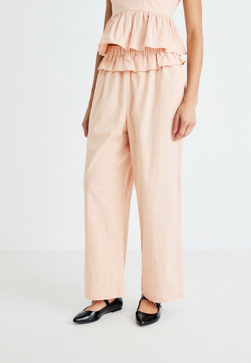 Conjunto de color melocotón que presenta una blusa de volantes en capas y pantalones anchos y holgados. El material parece suave; los zapatos son negros y tienen una correa.