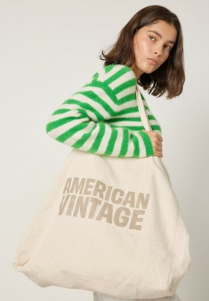Sac tote en toile crème avec un grand texte beige "AMERICAN VINTAGE" ; utilisateur portant un pull à rayures vertes et blanches, sac porté sur l'épaule.